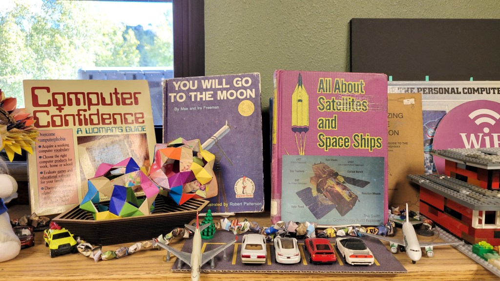 A desktop with older books including "Computer Confidence: A Woman's Guide", "You Will Go to the Moon" and more. Desk also includes small airplane and car tows and other ephemera. 